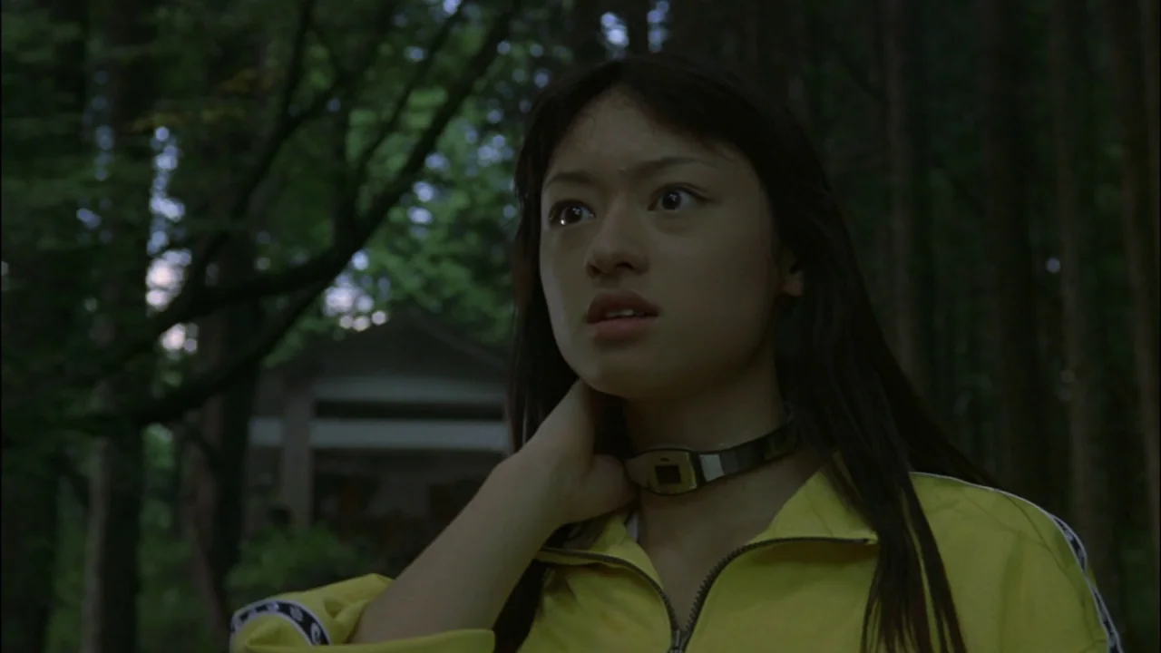 Battle Royale (2000) movie still: medium — A dense forest setting with a blurred structure visible in the background foliage. A young woman wears a…