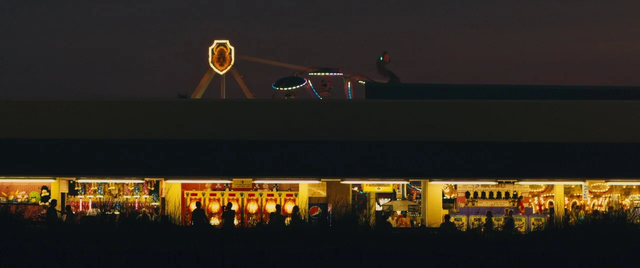 Blue Ruin (2013) movie still: establishing — A row of brightly lit carnival game stalls at night sits before a dark horizon. Silhouetted figures…