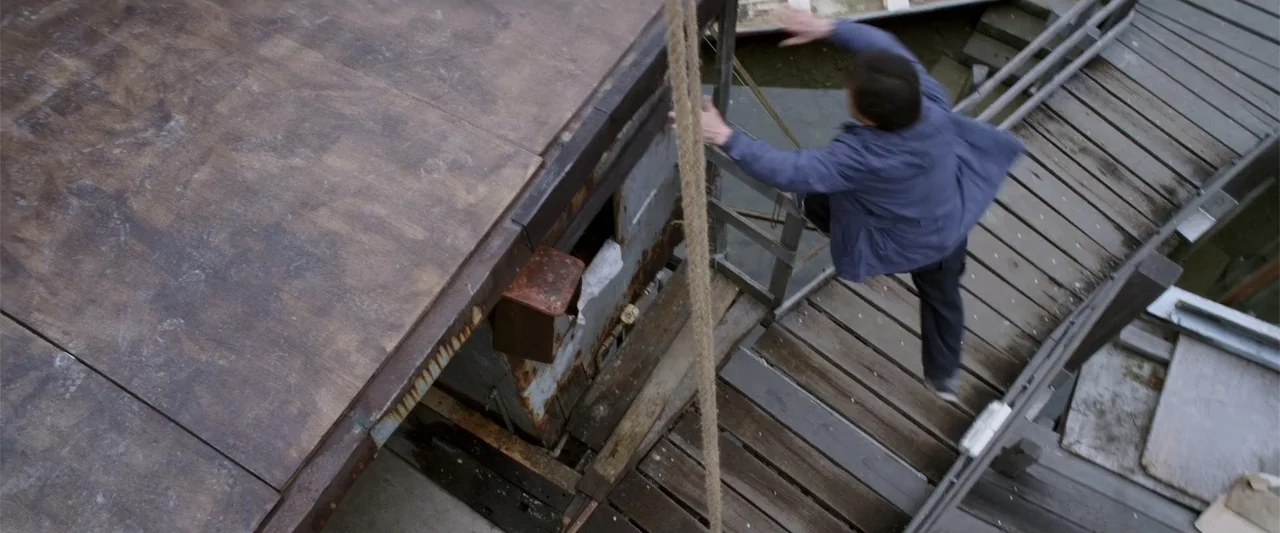 Skiptrace (2016) movie still: fullbody — A wooden pier and an adjacent boat deck are seen from a high angle. A man jumps from the boat deck onto the…