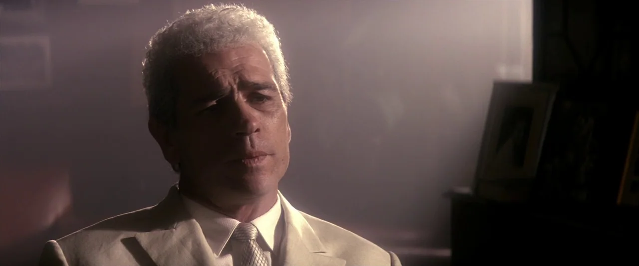 JFK (1991) movie still: closeup — A dimly lit room with hazy atmosphere and visible dark furniture in the background. A mature man in a white suit…