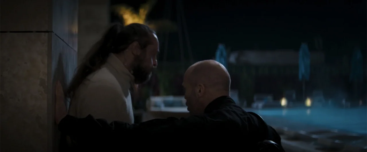 Operation Fortune: Ruse de Guerre (2023) movie still: medium — An outdoor area at night features a stone wall beside a lit pool. Two men stand close…