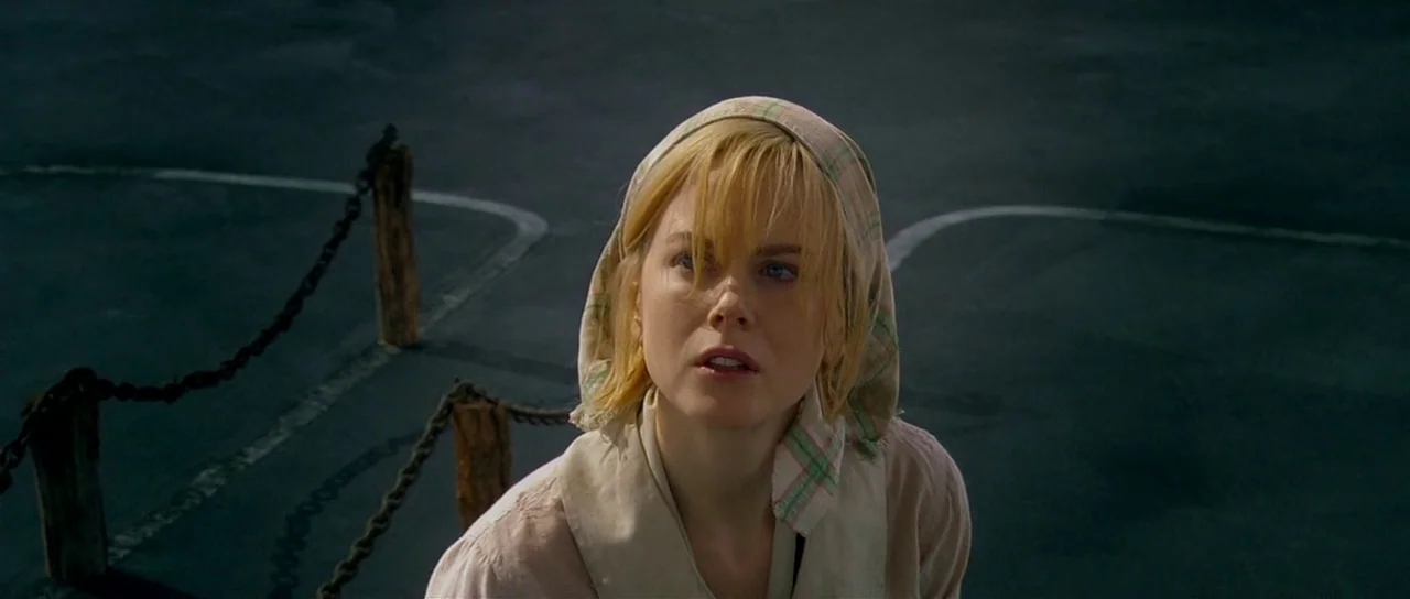 Dogville (2003) movie still: A young woman stands alone in an expansive, dark, and empty space, looking upward with a mixture of apprehension and…