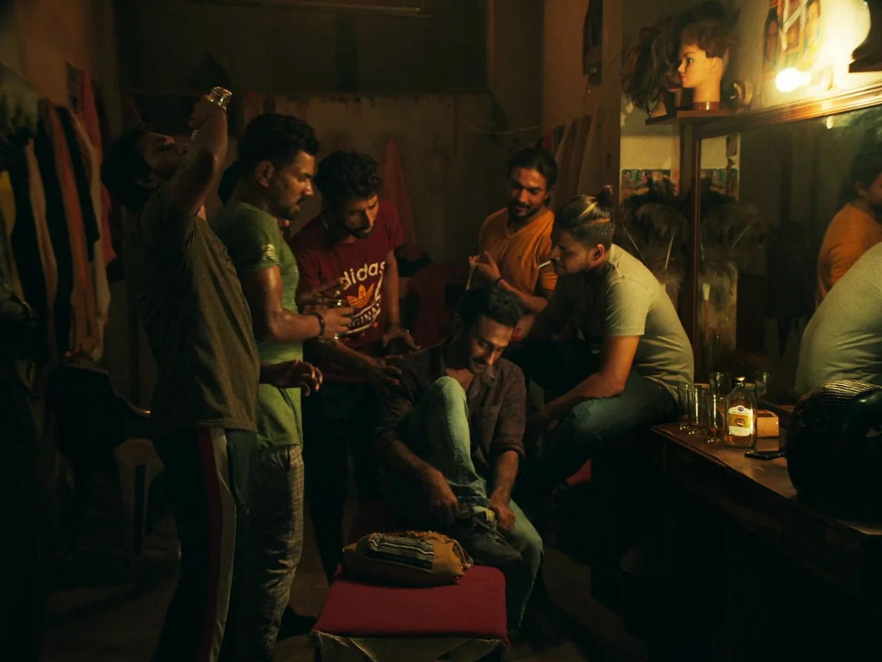 Joyland (2022) movie still: medium — A dimly lit room contains multiple men gathered around a central figure sitting down. The space is filled with…