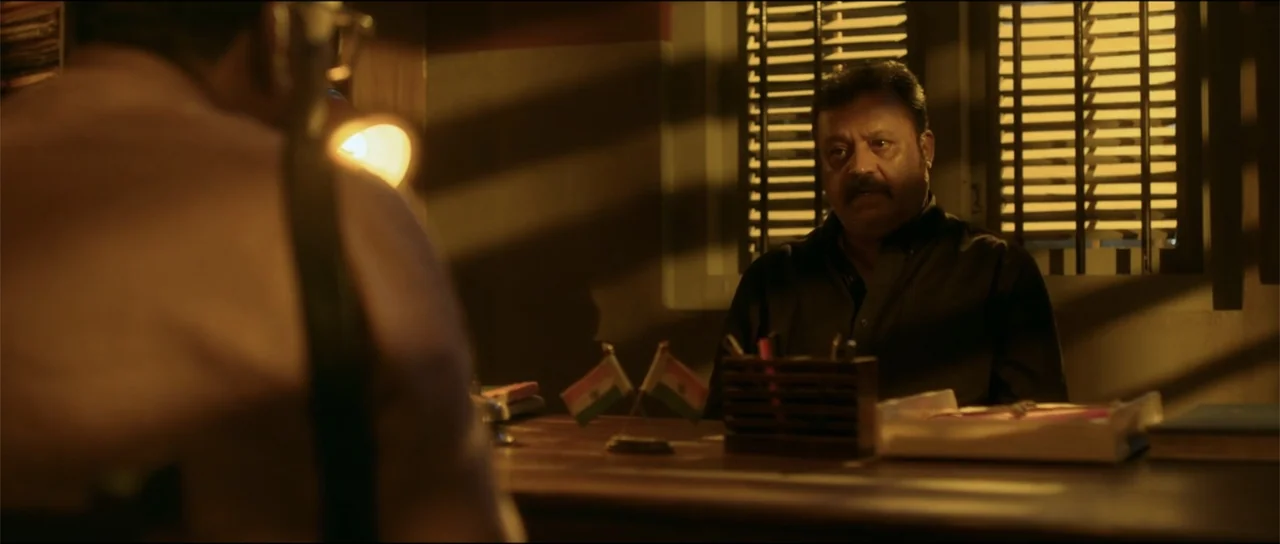 Garudan (2024) movie still: medium — An office interior features a wooden desk, venetian blinds, and small national flags. A man in a dark shirt…