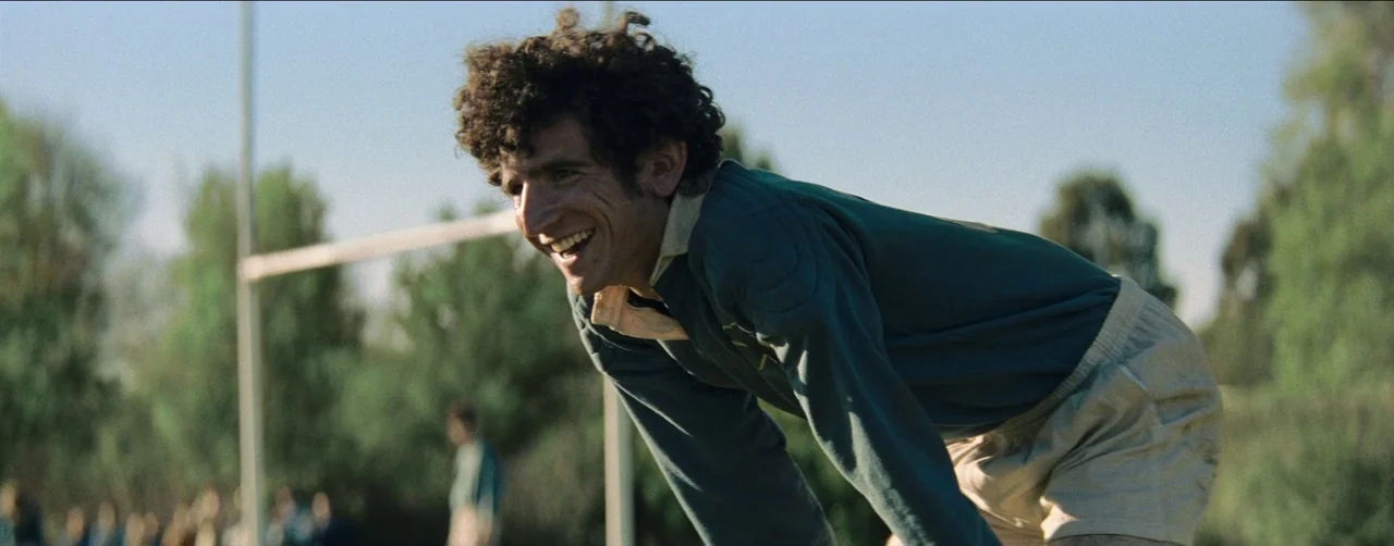 Society of the Snow (2023) movie still: medium — A rugby player stands on a grassy field with a metal goalpost in view. He leans forward with his…