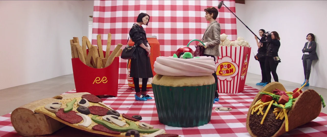 The Nowhere Inn (2020) movie still: wide — A sterile gallery space contains giant foam sculptures of fast food on a grid cloth. Two women face each…
