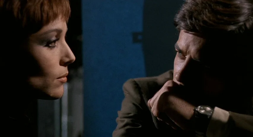 Perversion Story (1969) movie still: medium — A dimly lit room with a man and a woman in close proximity. The man sits with his hand on his chin…