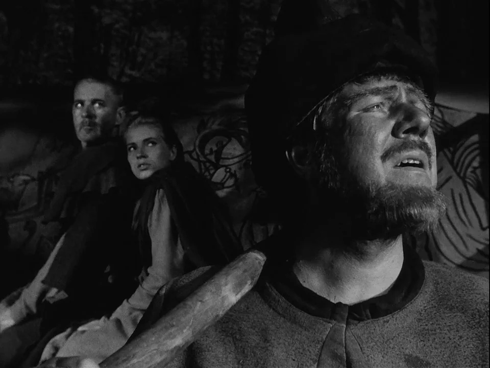The Seventh Seal (1957) movie still: medium — A dark interior space features rough, patterned walls behind three figures in medieval attire. The…