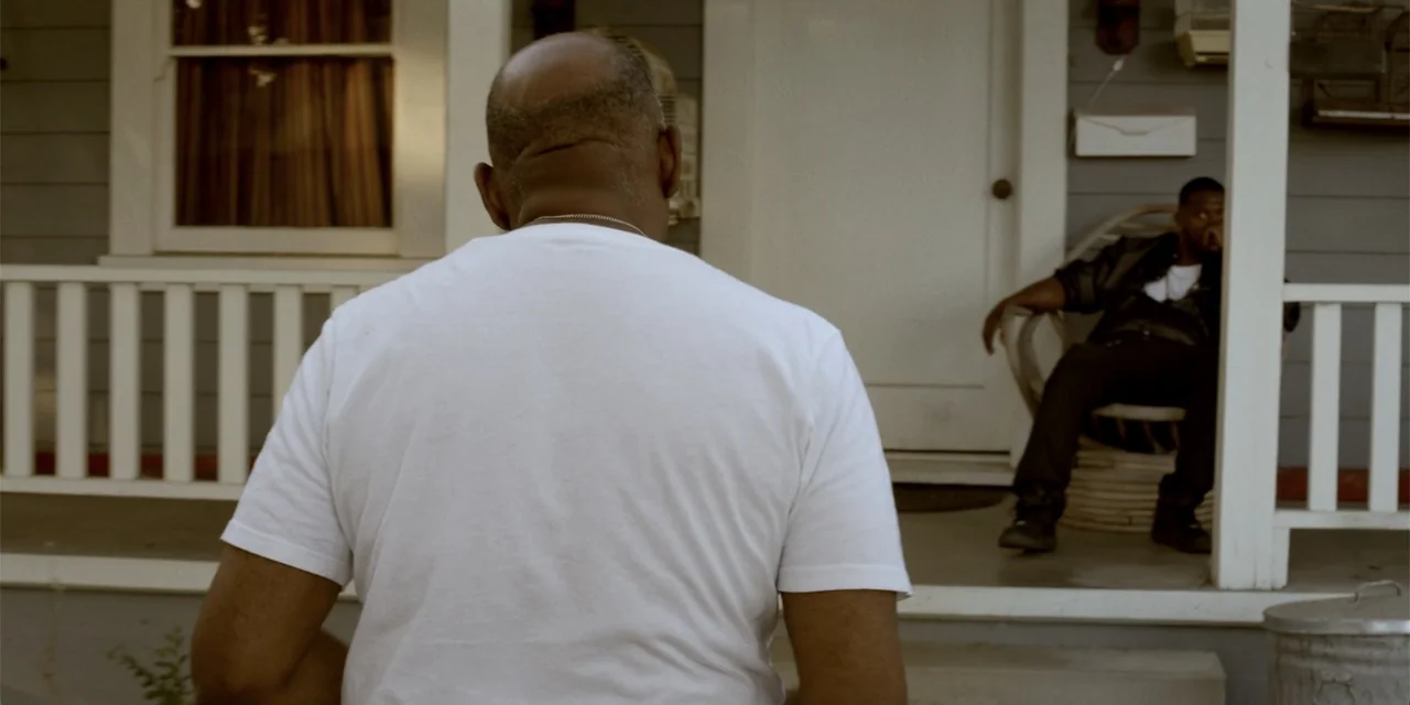 7th & Union (2021) movie still: medium — A residential house porch with grey wood siding and a white front door. One man stands facing the porch…