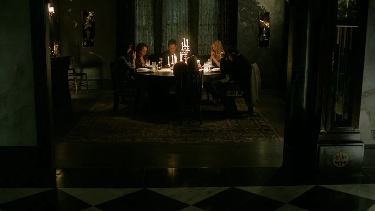Curse of Chucky (2013) movie still: wide — A dimly lit room contains a large round table surrounded by five seated people. The subjects sit in…