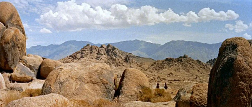 Ride Lonesome (1959) movie still: wide — A vast desert valley is framed by massive granite boulders and distant blue mountains. Two tiny silhouettes…