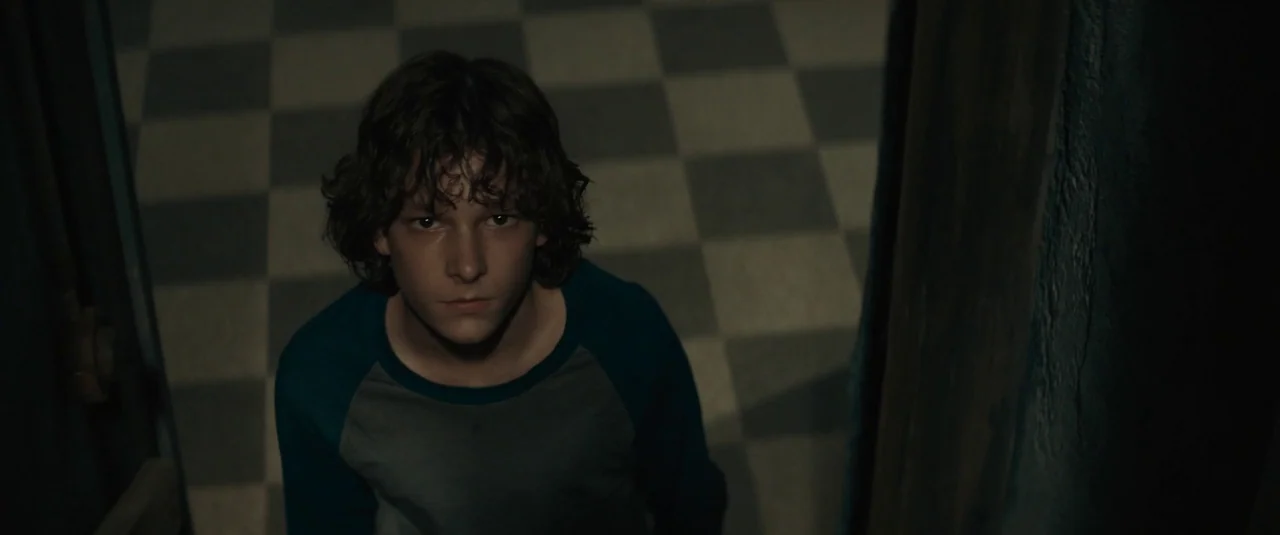The Black Phone (2021) movie still: medium — A stairwell features a checkered tile pattern on the floor surface. A teenage boy looks upward toward…