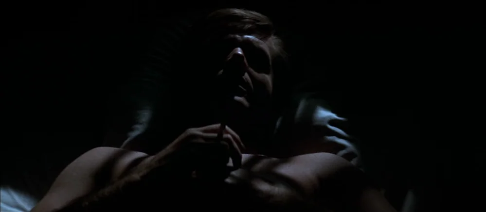 Carnal Knowledge (1971) movie still: closeup — A dark bedroom with white pillows visible behind a reclining male subject. The man lies on his back…