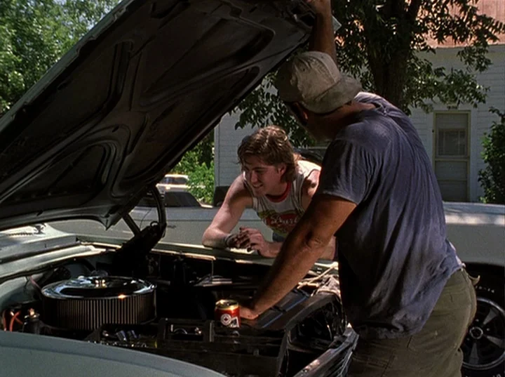 Slacker (1991) movie still: medium — A suburban residential driveway with a light blue vintage car parked under a tree. Two men lean over the open…