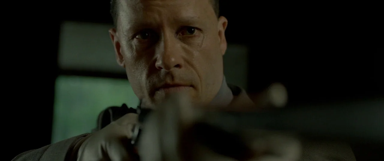 Lawless (2012) movie still: closeup — A man holds a firearm in a dark, dimly lit indoor setting. He stares directly ahead while aiming the weapon…