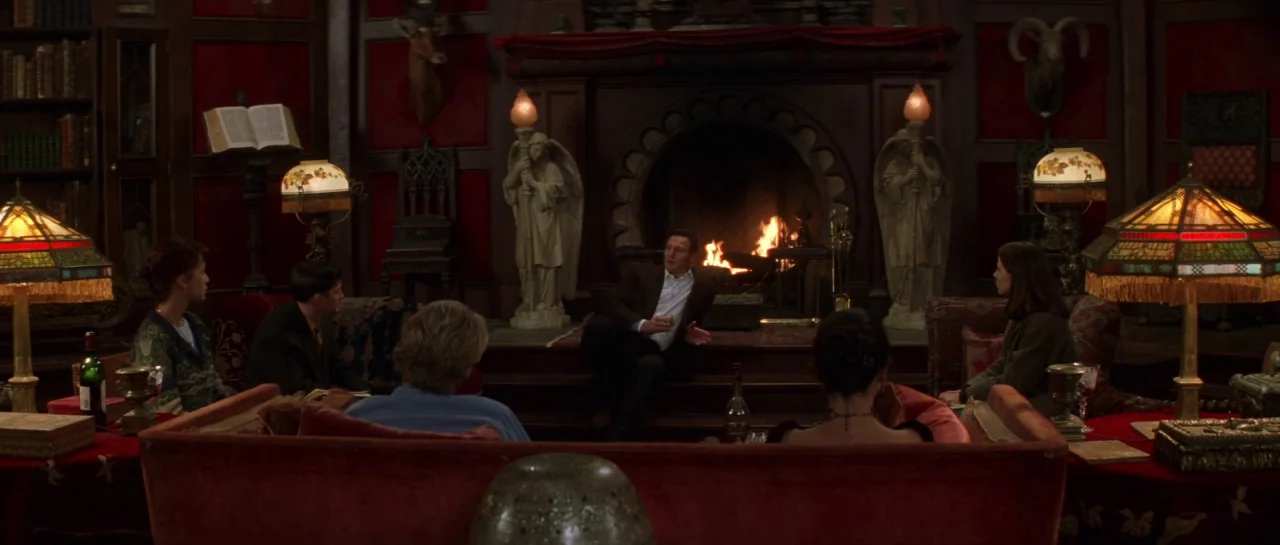 The Haunting (1999) movie still: wide — An opulent wood-paneled room features a large stone fireplace flanked by two angel statues. Five people sit…