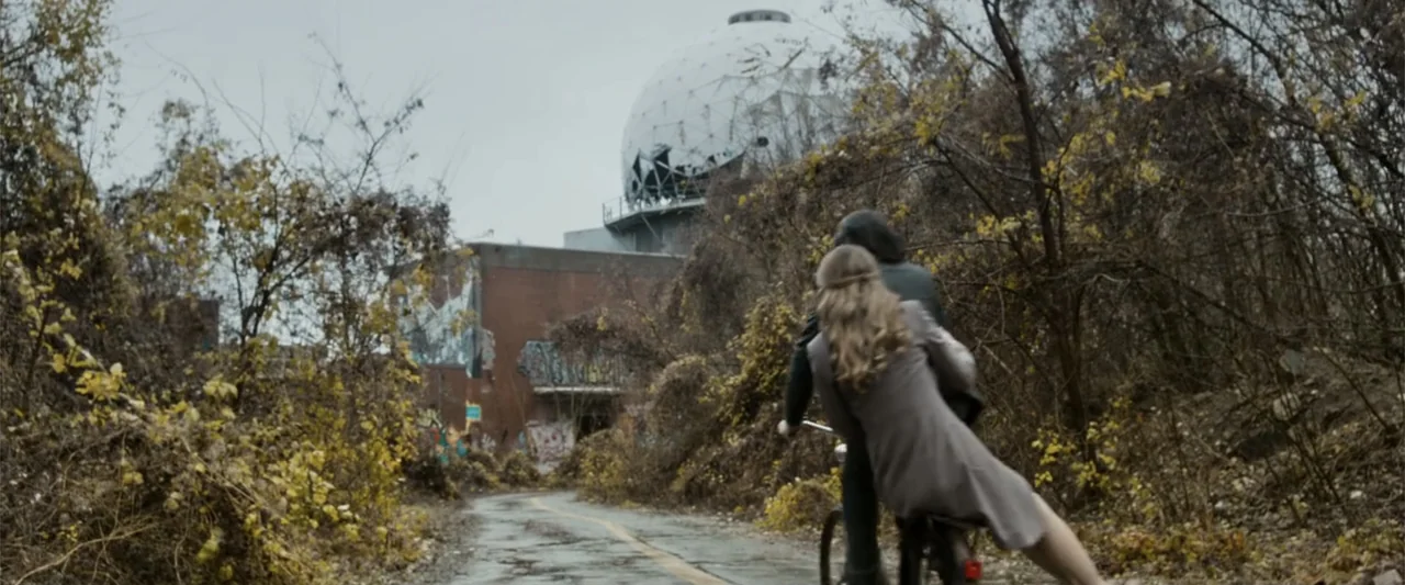 Heartburn (1986) movie still: fullbody — An abandoned road leads toward a brick building topped with a large radar dome. A woman sits on the back of…