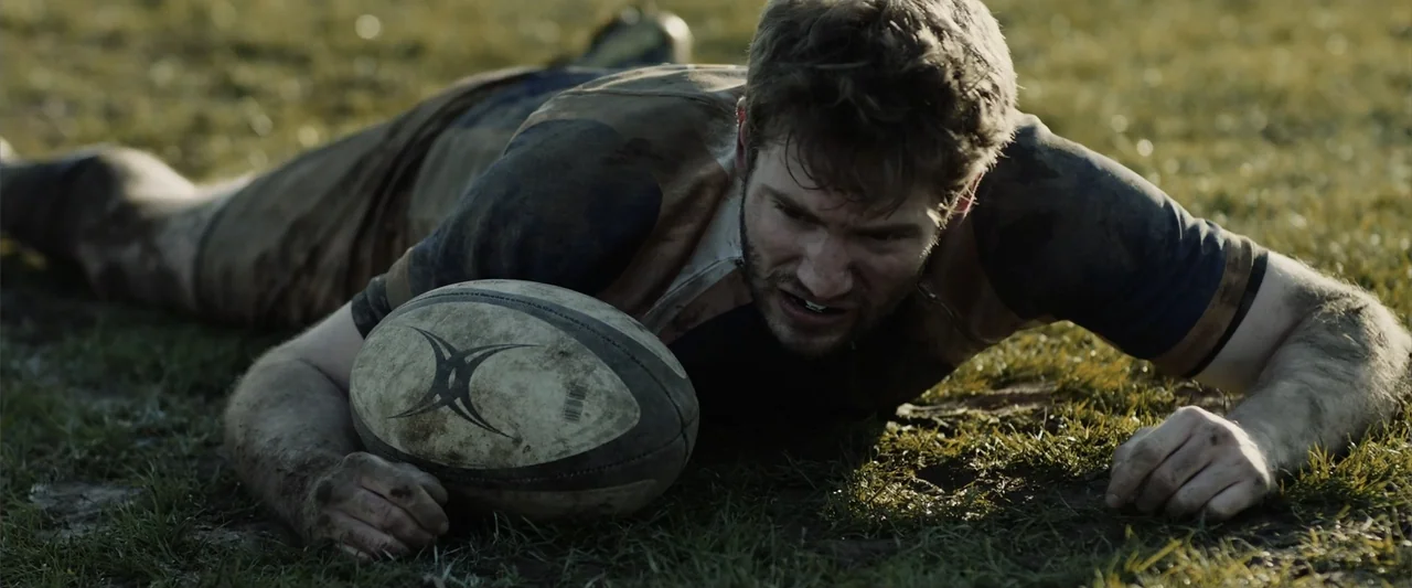 The Thorn On His Side (2014) movie still: medium — A muddy grass athletic field shows a player lying flat on the ground. A young man wearing a…