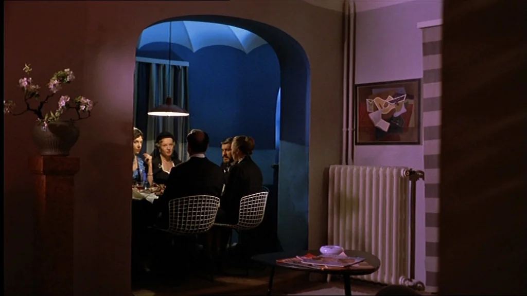 Lola (1981) movie still: wide — A modern interior space features a dining alcove with vibrant blue walls. Four people sit at a table while a…