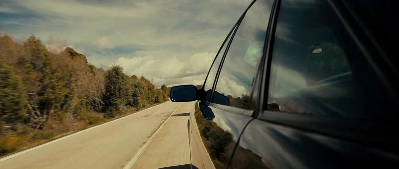 The Imposter (2012) movie still: wide — An asphalt road cuts through a rural landscape with sparse trees and cloudy skies. The side of a moving…