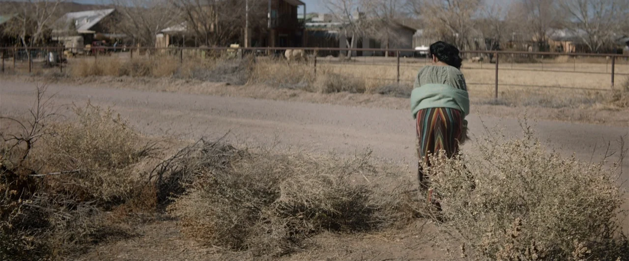 Cry Macho (2021) movie still: fullbody — A rural dirt road passes through a sparse landscape with arid brush. A woman walks away from the camera…