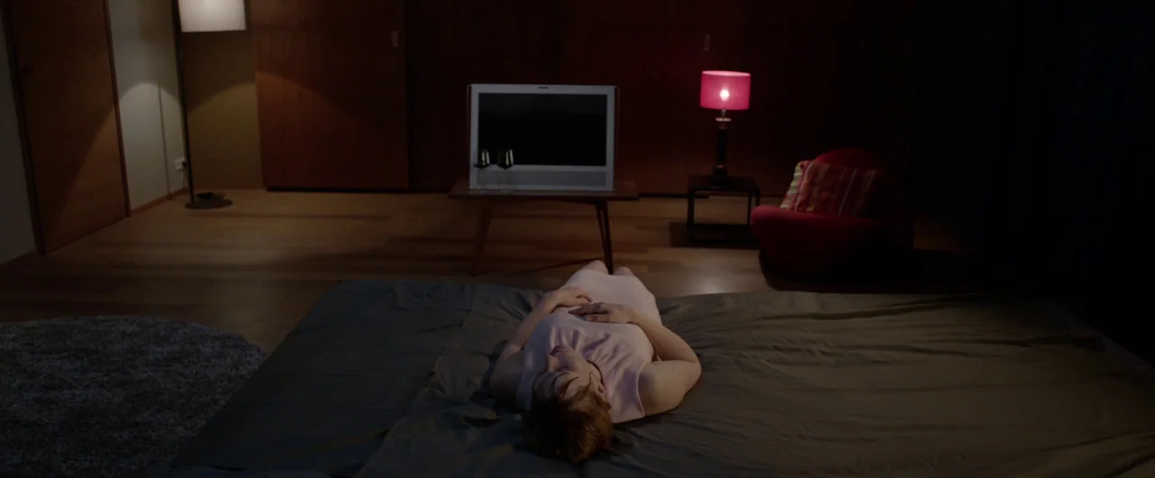Borgman (2013) movie still: wide — A dark hotel room features dark wooden panels, a flat-screen television, and modern red furniture. A woman lies…
