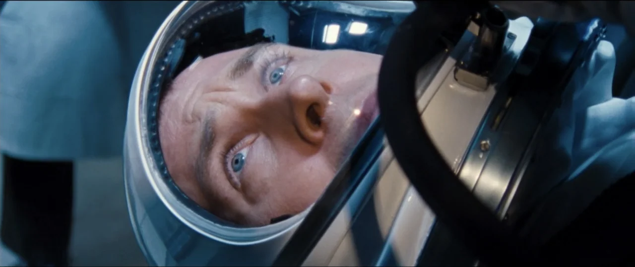 First Man (2018) movie still: closeup — A sterile interior environment contains a man encased in a metal-rimmed space helmet. The subject stares…