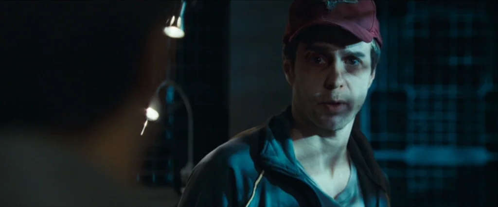 Moon (2009) movie still: medium — A man wearing a baseball cap stands in a dimly lit dark industrial environment. The subject directs a gaze toward…