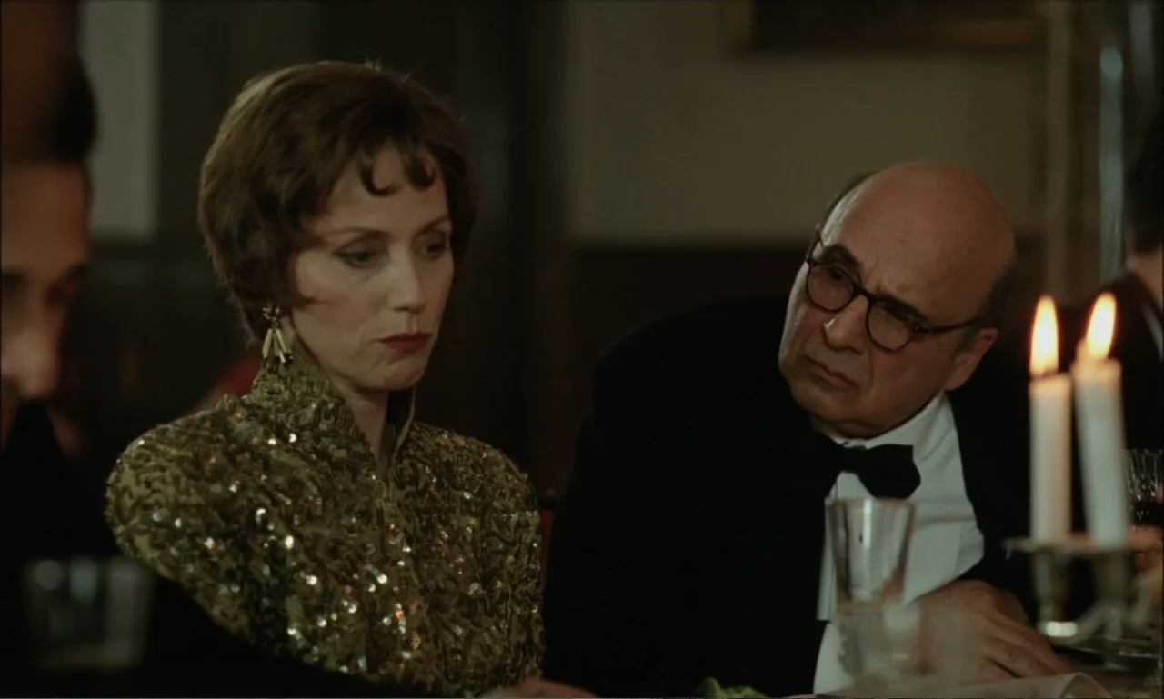 The Brutalist (2024) movie still: medium — A dimly lit formal dining room features a table set with candles. A woman in a sequined dress sits next…