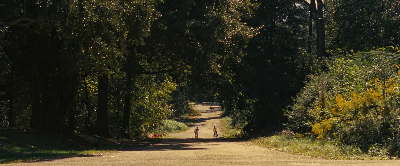 All Dirt Roads Taste of Salt (2023) movie still: wide — A long, straight paved road cuts through a dense canopy of tall, green trees. Two children…