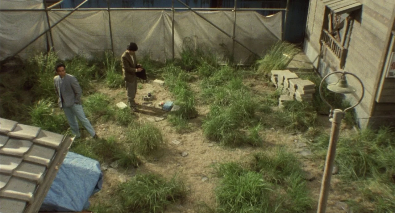 The Discarnates (1988) movie still: wide — An overgrown vacant lot is enclosed by white scaffolding fabric and a wooden wall. Two men in suits…