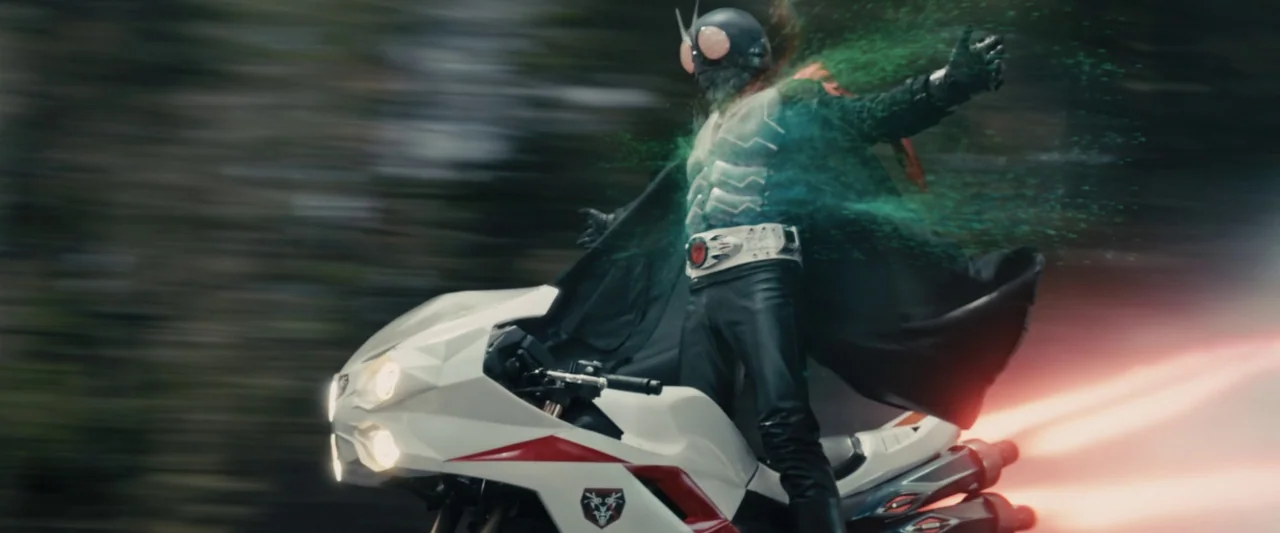 Shin Kamen Rider (2023) movie still: medium — A masked hero rides a white sportbike on a blurred road in daylight. Green digital particles dissipate…
