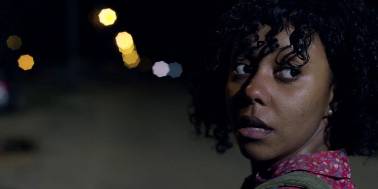 Jakob’s Wife (2021) movie still: closeup — A dark outdoor urban setting at night with out-of-focus street lights in the background. A woman looks…