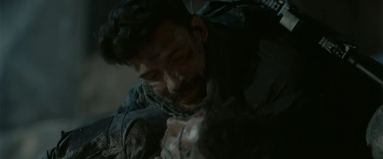 Hounds of War (2024) movie still: closeup — A dark, dirt-covered ground serves as the backdrop for two men in tactical gear. One man cradles the…