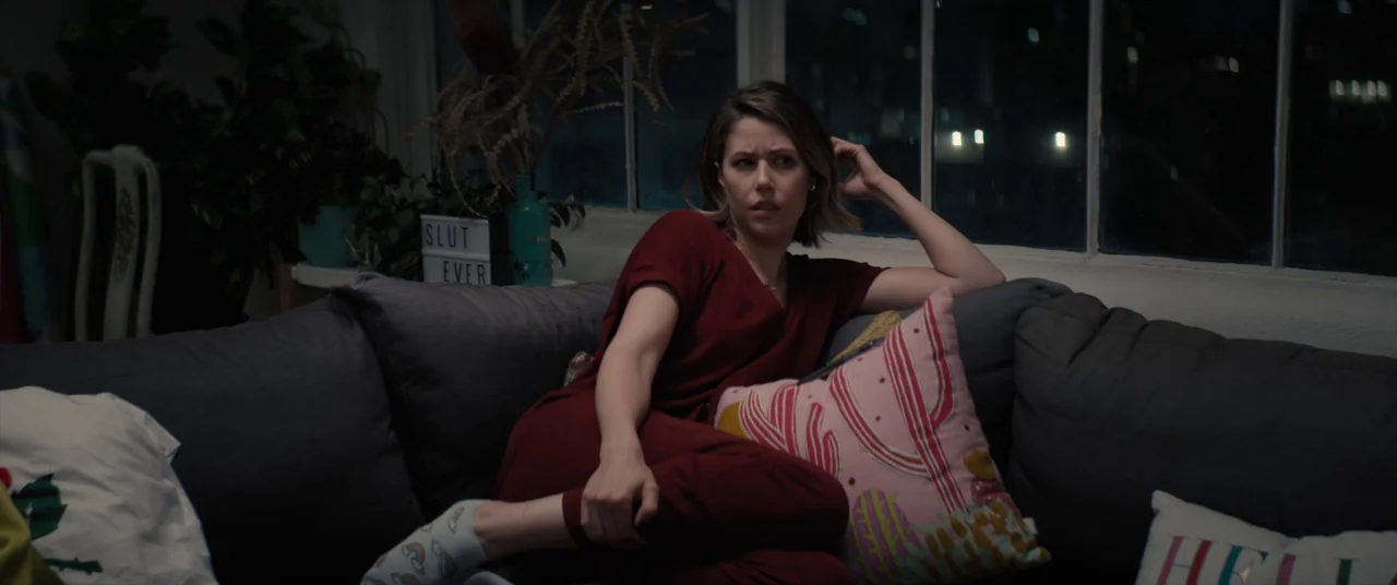 Tone Deaf (2019) movie still: medium — A modern apartment living room features a dark gray sofa against a multi-pane window at night. A young woman…