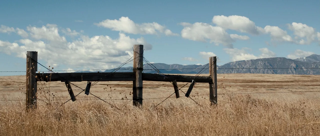 Cop Car (2015) movie still: establishing — A wooden barbed wire fence divides a flat, dry, golden-brown prairie landscape. Rolling mountains rise…