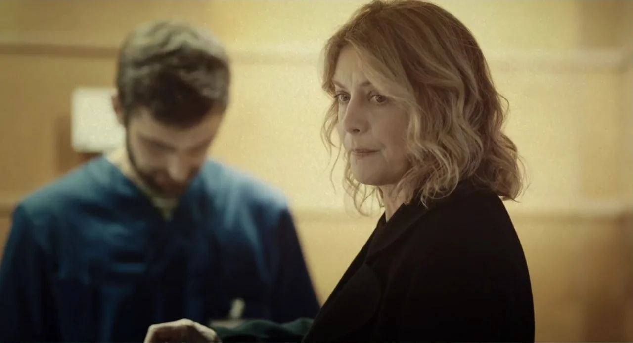 Elles (2018) movie still: medium — A light-colored institutional hallway features textured walls and fluorescent lighting. A woman in a dark coat…