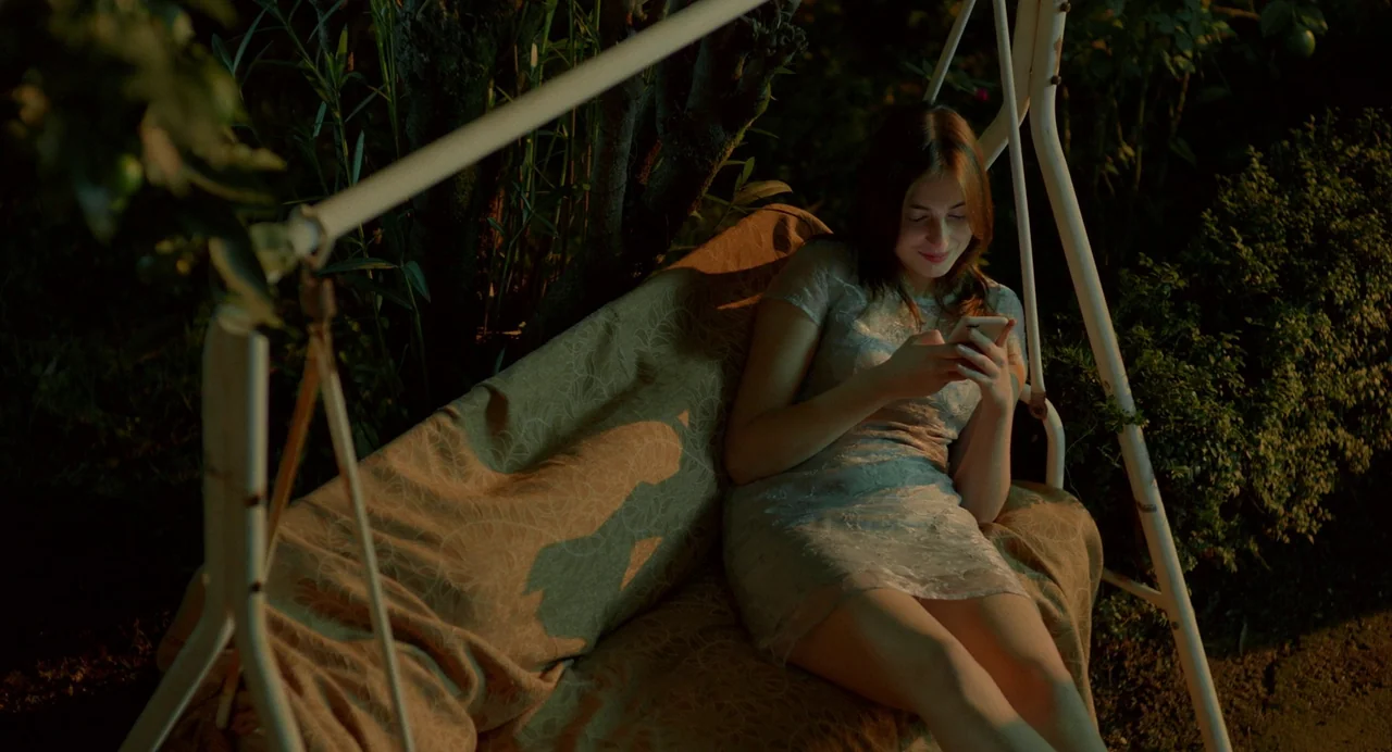Blackbird Blackbird Blackberry (2023) movie still: medium — A woman sits on a metal porch swing at night surrounded by foliage. She focuses on her…