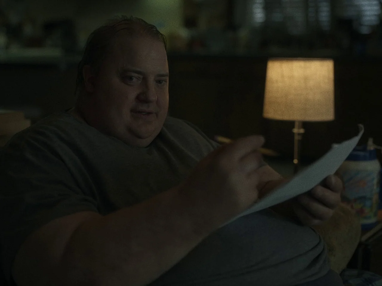 The Whale (2022) movie still: medium — A dark, dimly lit room contains a person sitting next to a glowing table lamp. A man holds a sheet of paper…