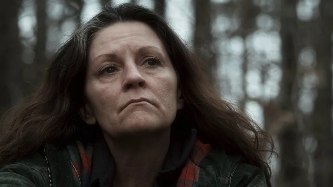 Winter’s Bone (2010) movie still: closeup — A woman stands in a dense, out-of-focus wooded area during a cloudy day. She looks upward with a…