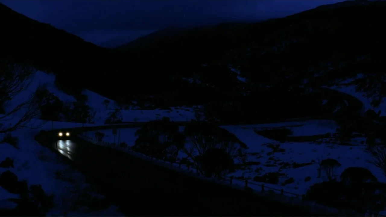 Emily (1976) movie still: wide — A dark, winding road passes through a snow-covered mountain valley under a deep blue sky. A distant vehicle travels…