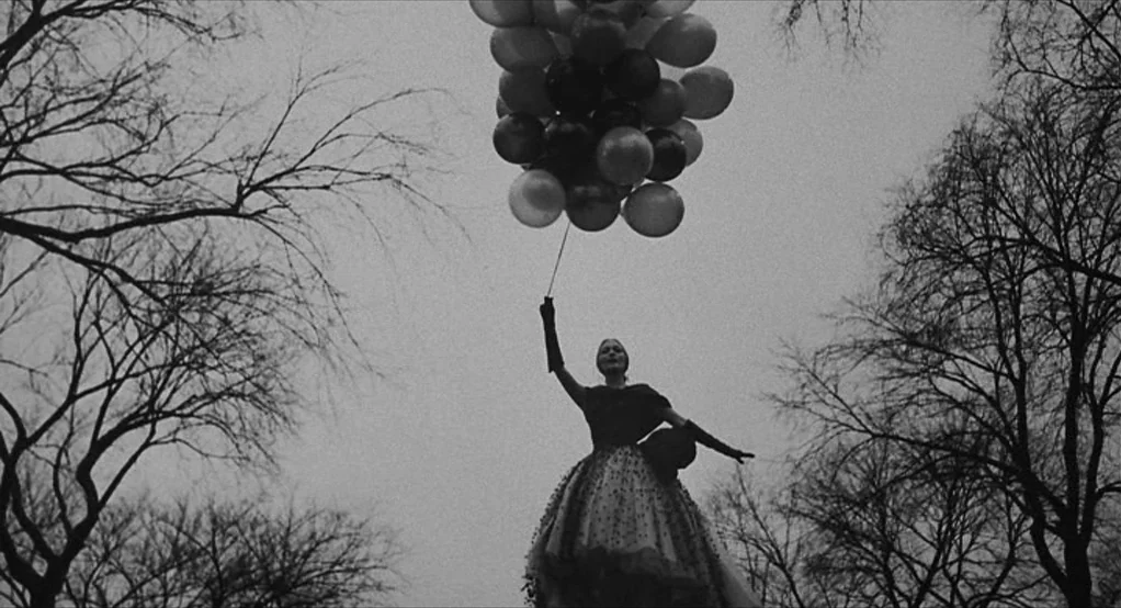 Puzzle of a Downfall Child (1970) movie still: fullbody — A woman stands in a leafless park under a grey sky. She holds a large cluster of balloons…