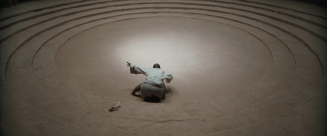Dune: Part 2 (2024) movie still: wide — A wide, circular sand pit features concentric ring patterns pressed into the terrain. A man in a white gi…