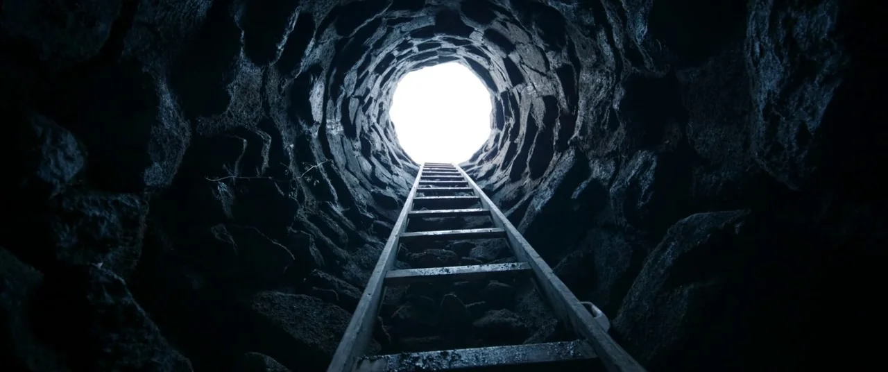 A Hidden Life (2019) movie still: establishing — A deep circular well shaft is built from rough stacked dark basalt stones. A wooden ladder extends…