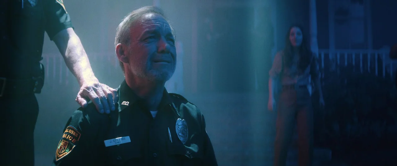 No One Will Save You (2023) movie still: medium — An older police chief kneels on a dark porch bathed in cool blue light. A hand rests on his…