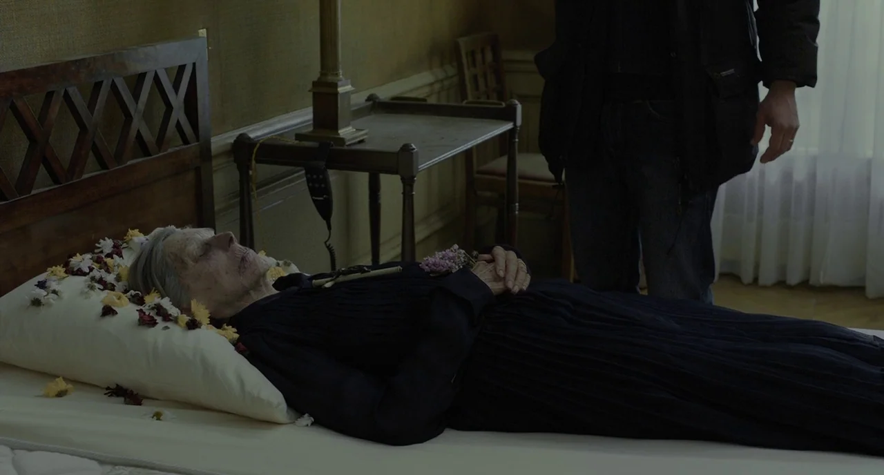 Amour (2012) movie still: medium — A sparsely furnished room contains a woman lying on a bed with scattered flowers. A standing man wearing a winter…