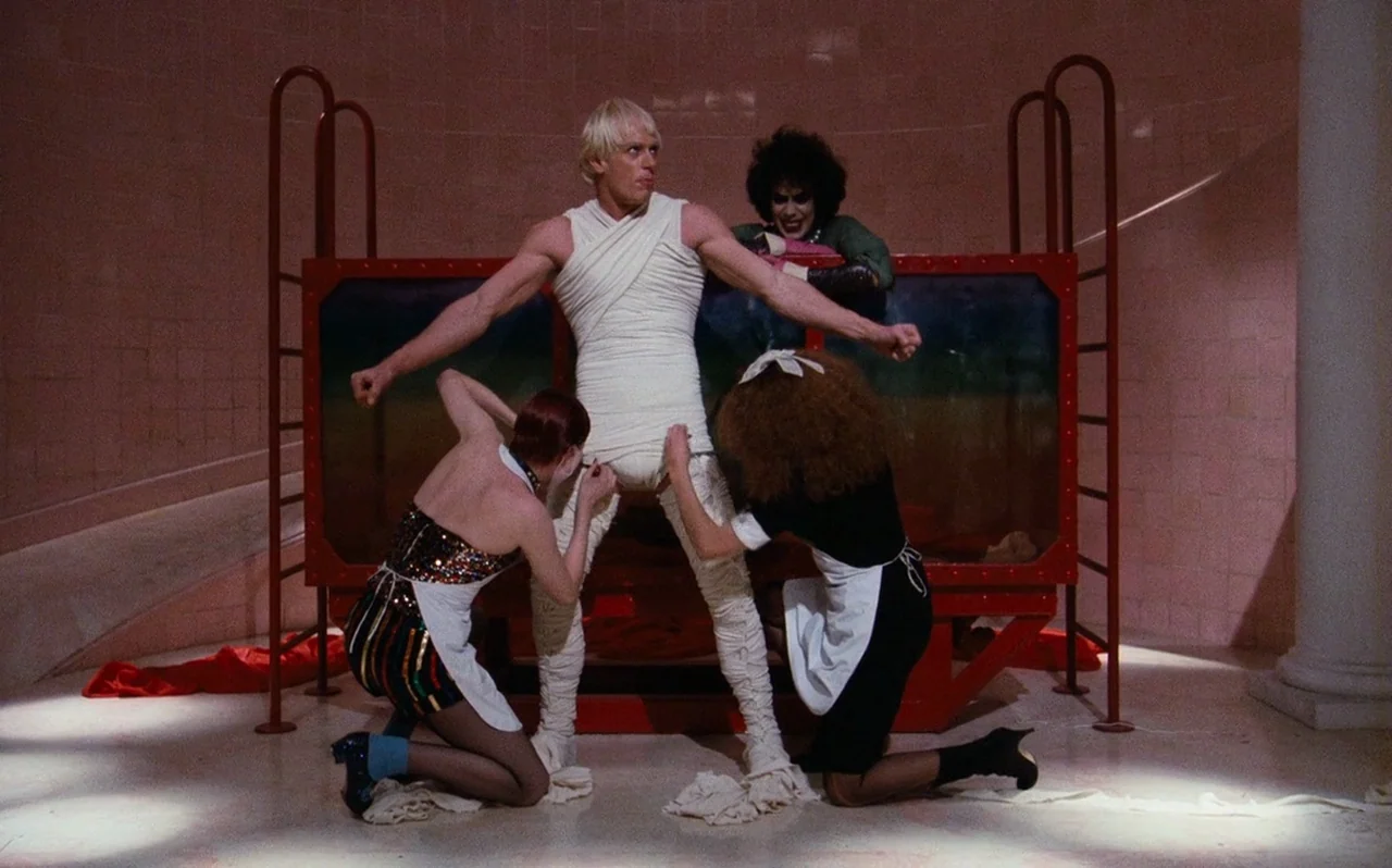 The Rocky Horror Picture Show (1975) movie still: fullbody — A pink tiled laboratory room with a large red-framed glass tank in the center. Two…