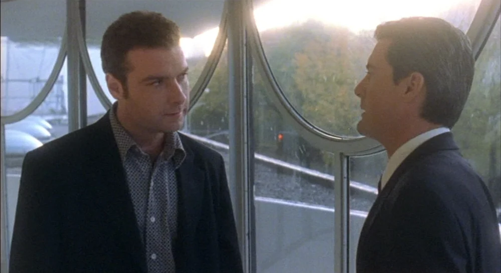 Hamlet (2000) movie still: medium — An interior hallway with circular glass panels overlooks trees during a bright day. Two men in formal business…