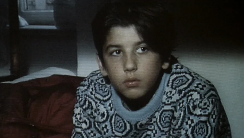 71 Fragments of a Chronology of Chance (1994) movie still: closeup — A dark indoor space with a boy seated in front of a wall. The boy is looking…