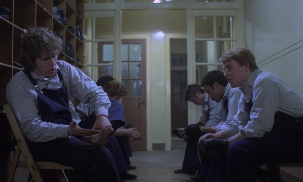 Scum (1979) movie still: medium — A locker room with wooden cubbies along one wall and a view through glass doors. A group of boys sits on benches…